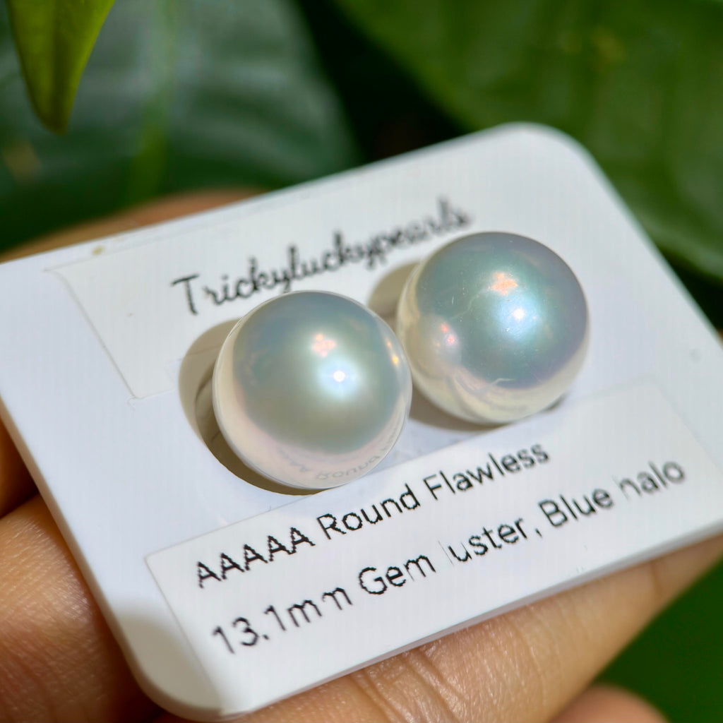 A close-up of two round iridescent freshwater pearls on a white display card held by a hand, with green leaves in the background. The card reads 'TrickyLuckyPearls' and 'AAAAA Round Flawless 13.