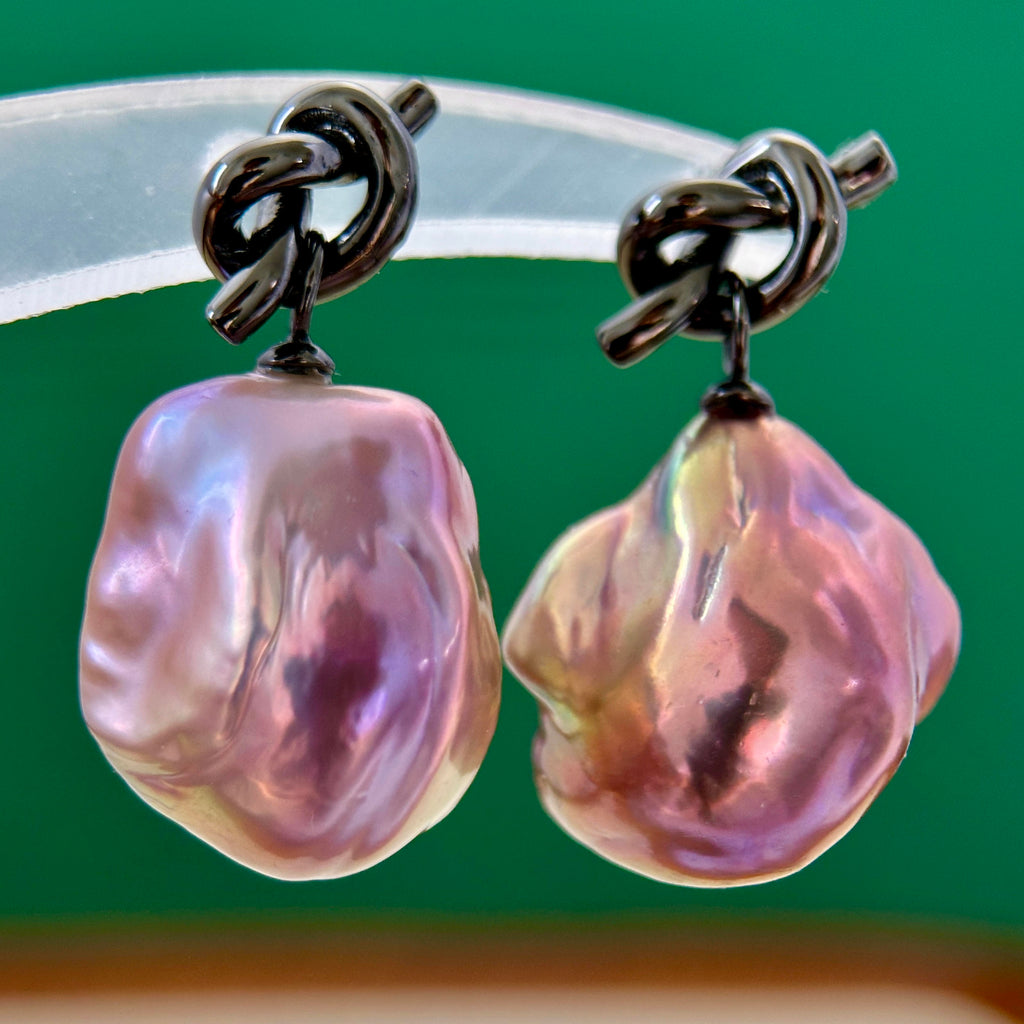 A pair of baroque pearl drop earrings with dark metal knotted posts, displayed on a clear acrylic stand against a solid green background.