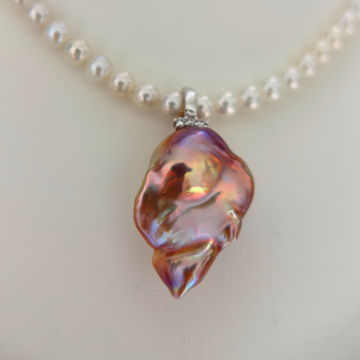 A close-up of a pearl necklace featuring a large, organic-shaped iridescent baroque pearl pendant in pink, purple, and gold tones, suspended from a silver-toned chain with small clear pearl.