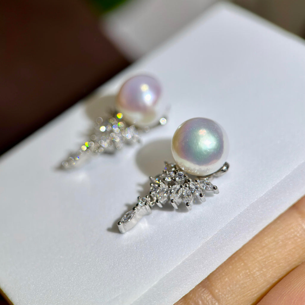 A pair of silver-toned drop earrings with round iridescent white pearls and sparkling clear stones, displayed on a white card with a finger holding the corner.