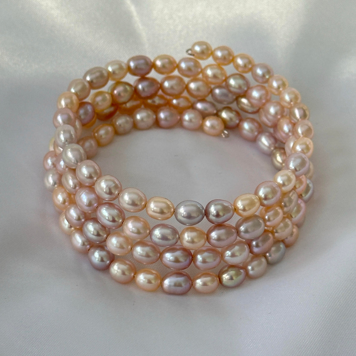 A multi-strand baroque pearl bracelet in soft peach, pink, and champagne tones, displayed on a white satin fabric background.
