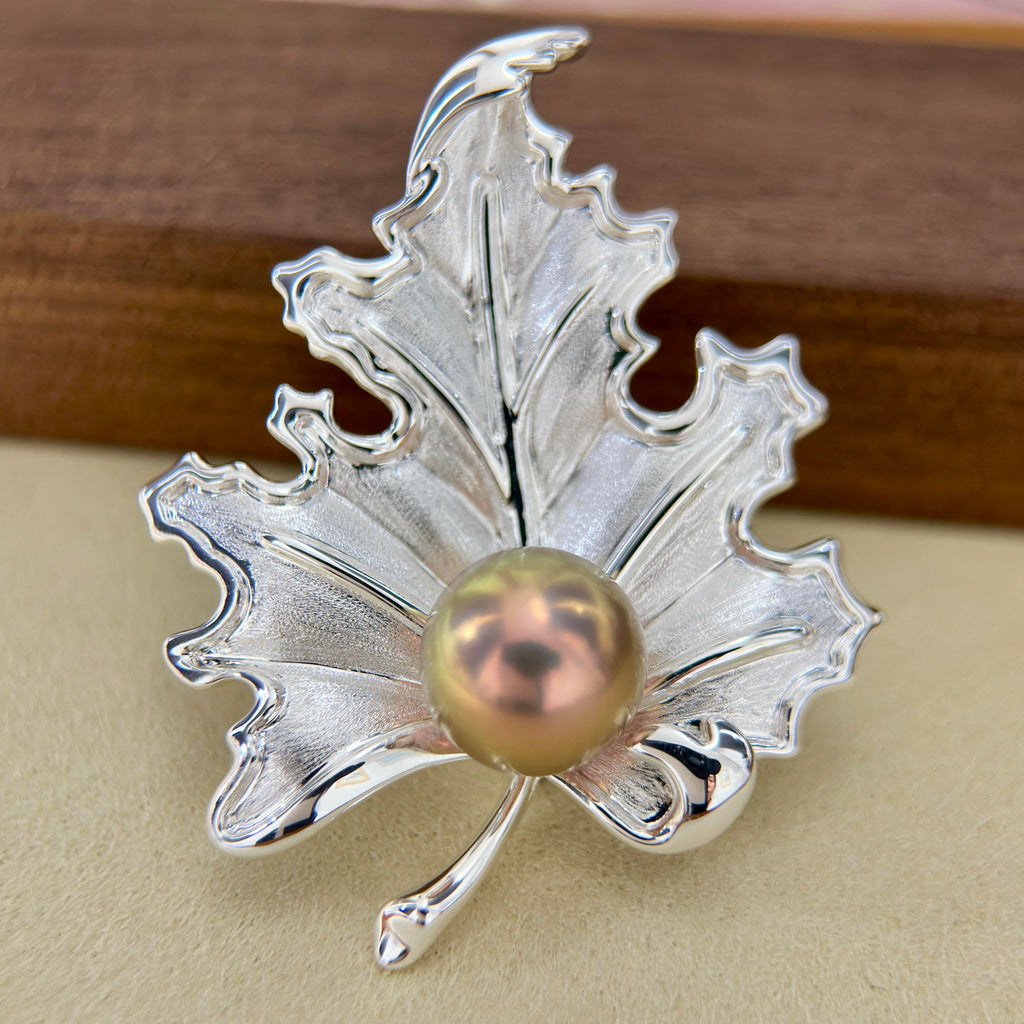 A silver maple leaf-shaped brooch or pin with a central iridescent pearl, photographed close-up on a beige surface with a blurred brown wooden background.