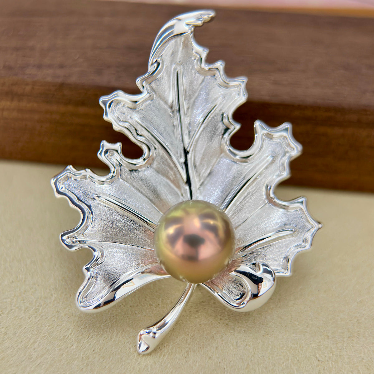 A silver maple leaf-shaped brooch or pin with a central iridescent pearl, photographed close-up on a beige surface with a blurred brown wooden background.