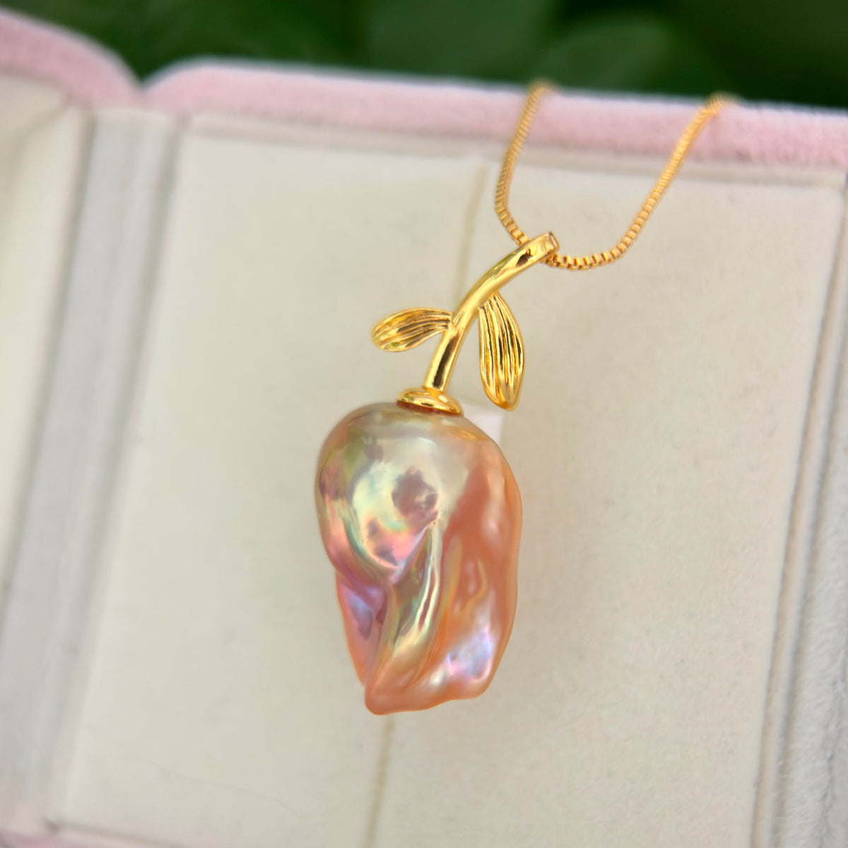 A gold pendant necklace featuring a large, irregular baroque pearl shaped like a peach or mango, resting on a soft cream-colored jewelry box.