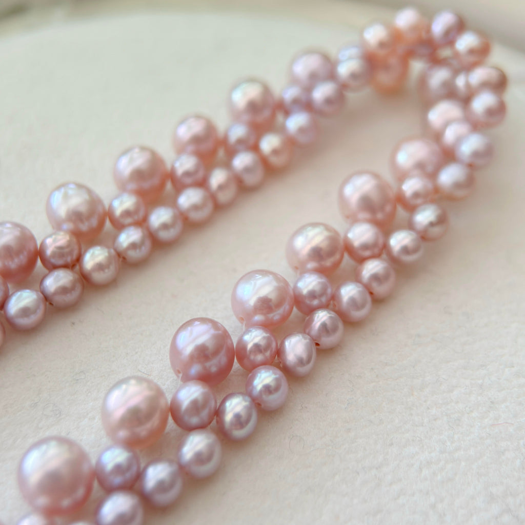 A close-up of a strand of pink freshwater pearls arranged in two rows on a light beige textured surface.