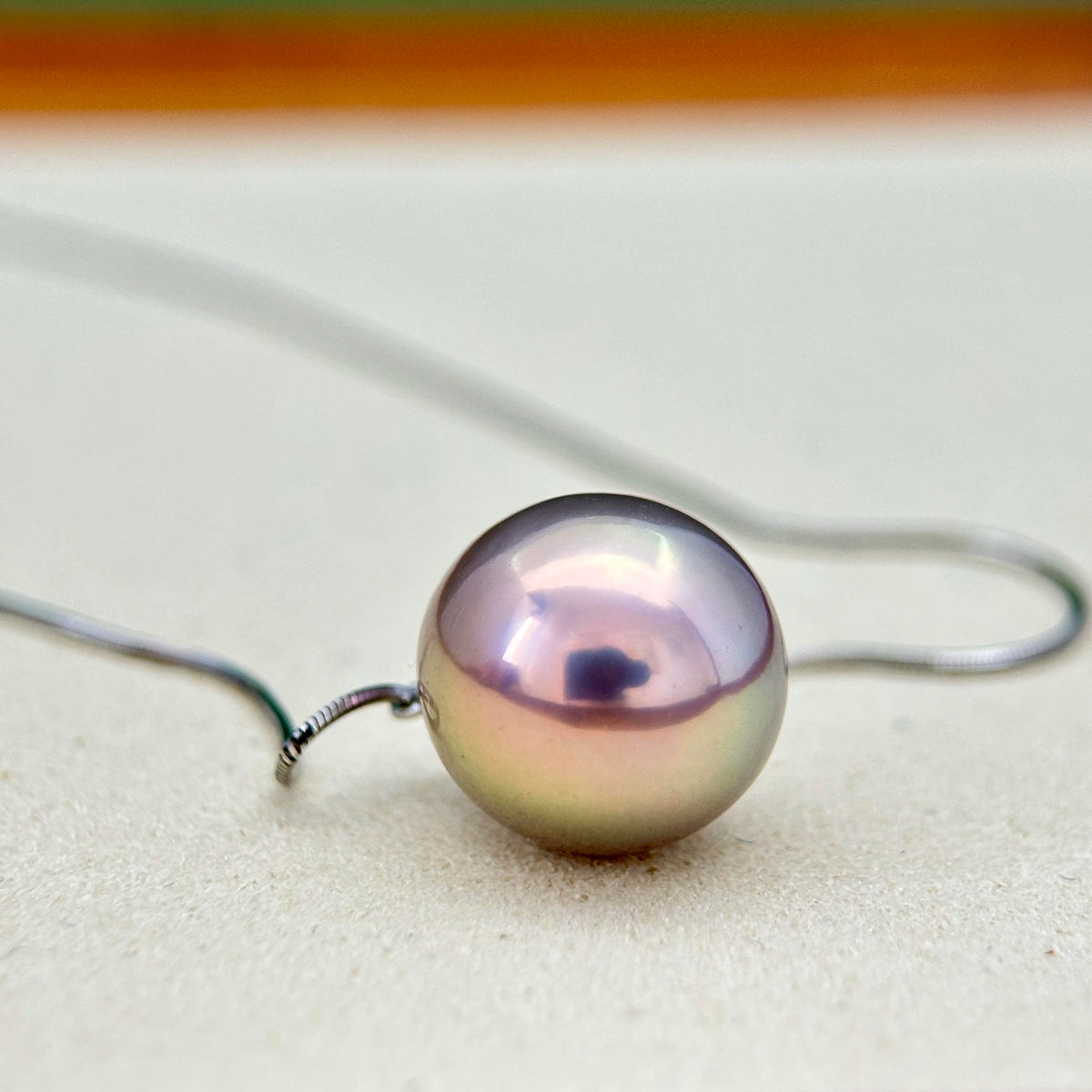 A single round iridescent pearl pendant on a thin silver wire necklace, resting on a light beige textured surface.