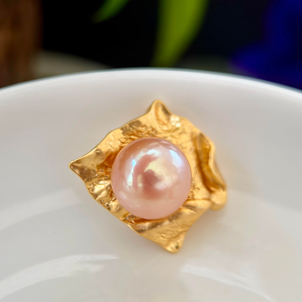 A gold pendant featuring a textured, leaf-like setting holding a round, lustrous pink pearl, resting on a white ceramic surface.