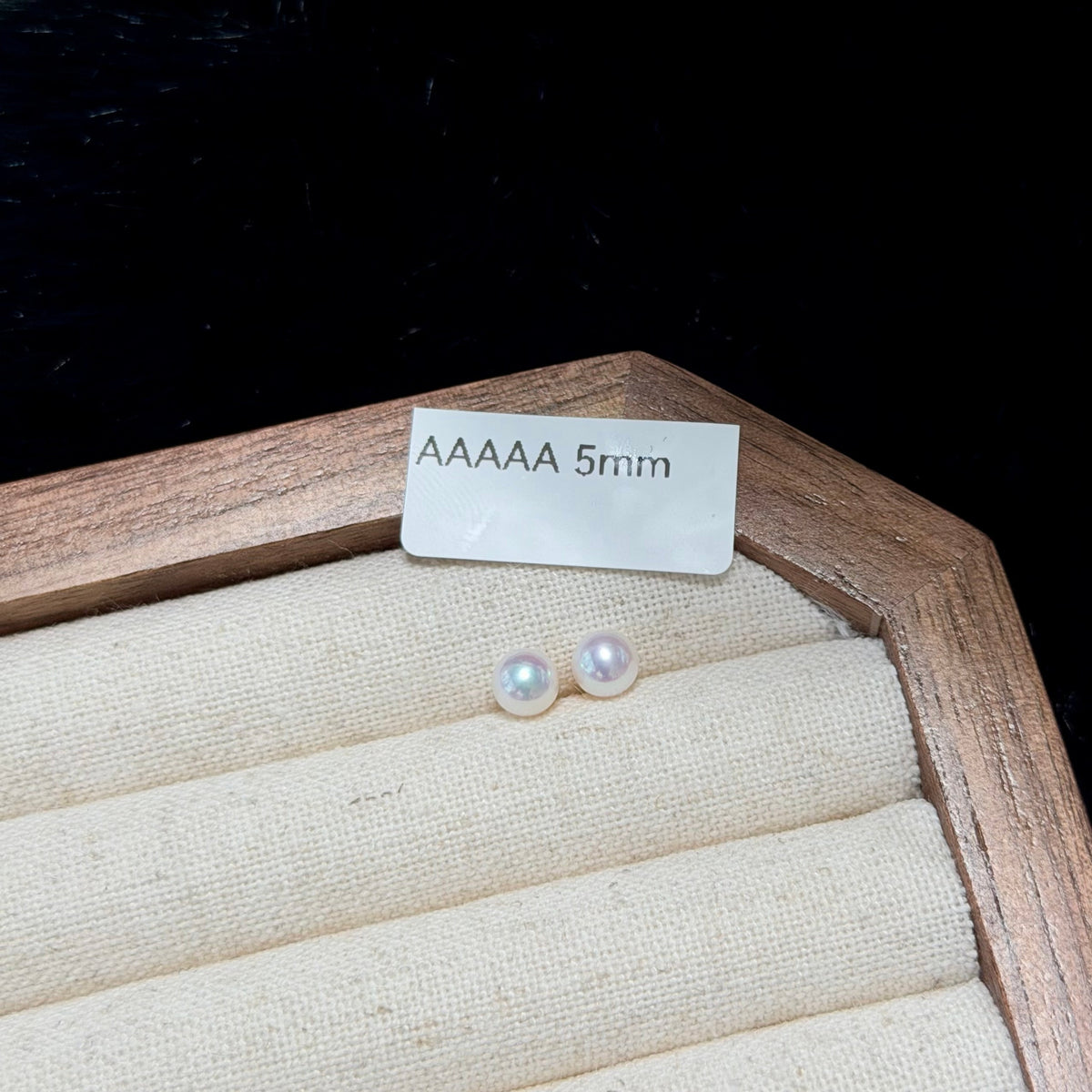A pair of small round white pearl stud earrings displayed on a beige fabric-lined jewelry box, with a small white label reading 'AAAAA 5mm' above them against a black background.