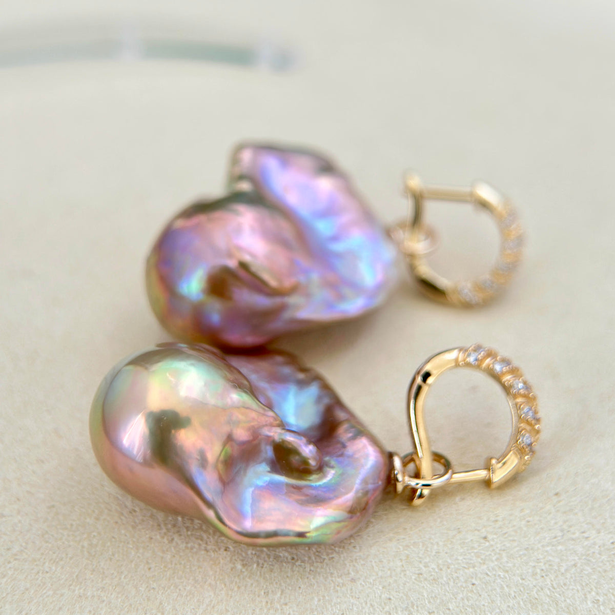A pair of gold hoop earrings with large, baroque-shaped iridescent pearl drops. The pearls exhibit shimmering hues of pink, purple, and green, and are attached to gold hoops embellished with small clear stones.