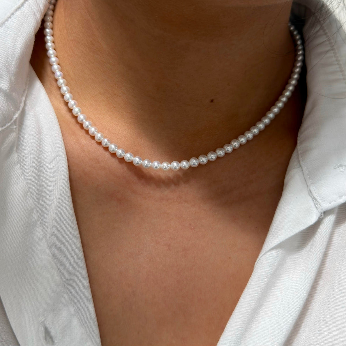 A close-up of a person wearing a delicate white pearl choker necklace with a white collared shirt.