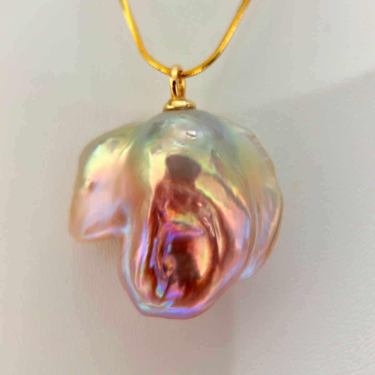 A gold chain necklace featuring a large, irregular baroque pearl pendant with iridescent pink, purple, and green hues, shown against a plain light background.