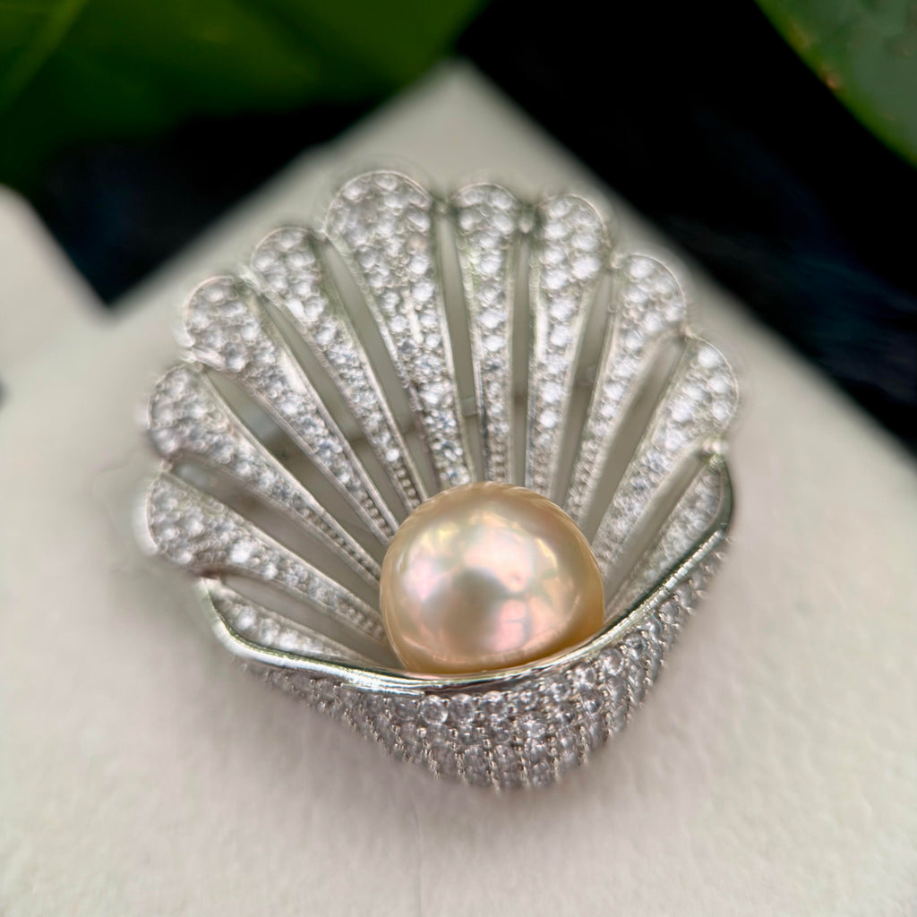 A scallop-shaped silver ring featuring a central freshwater pearl with iridescent pinkish-gold tones, surrounded by pavé-set clear stones, resting on a white textured surface.