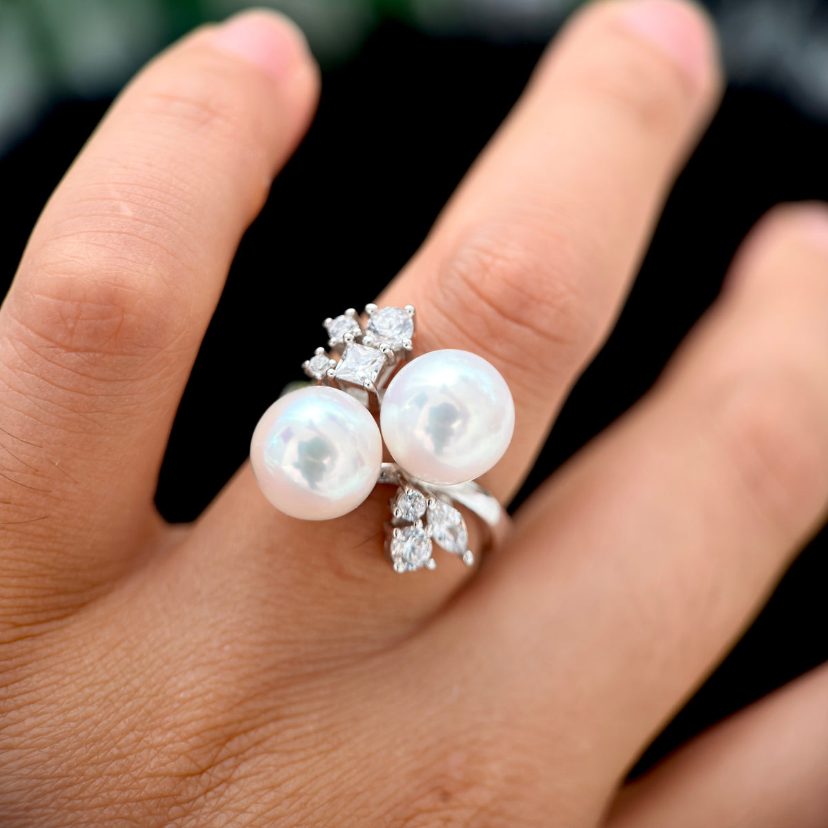 A close-up of a hand wearing a silver ring with two large white pearls and small clear stones in a leaf-like cluster setting.