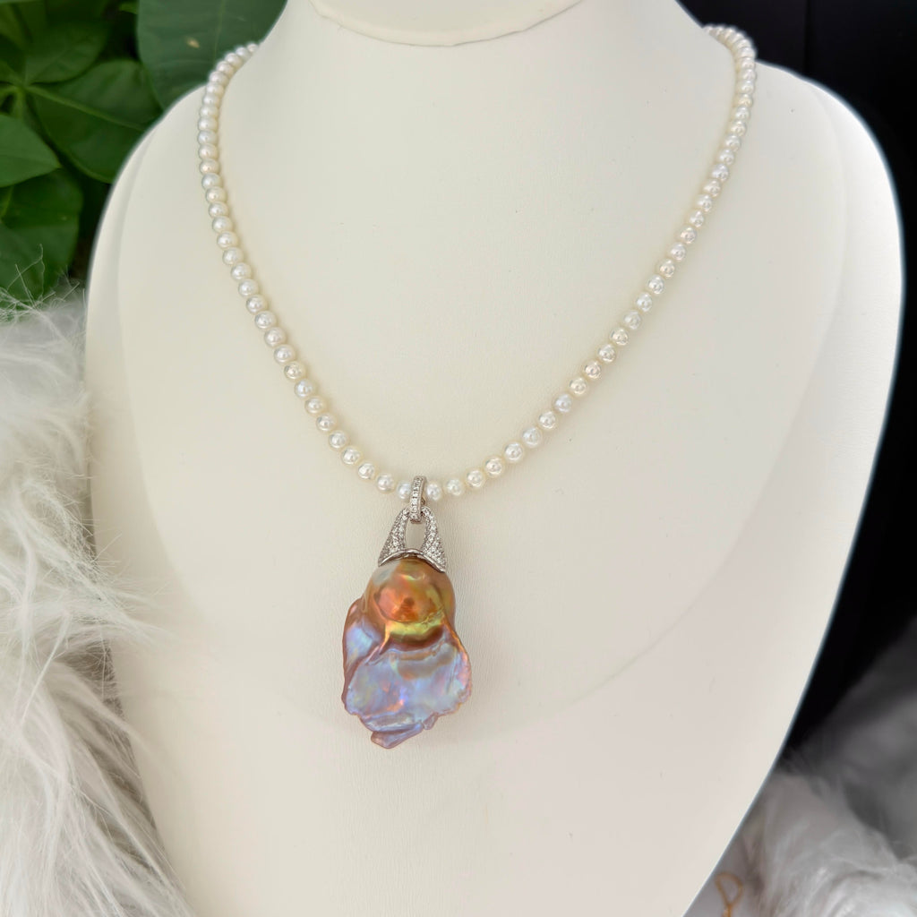 A pearl necklace with a large baroque shell pendant displayed on a white mannequin bust. The necklace features small, round white pearls and a silver-toned bezel holding a vibrant, iridescent shell with hues of orange, green, and blue.