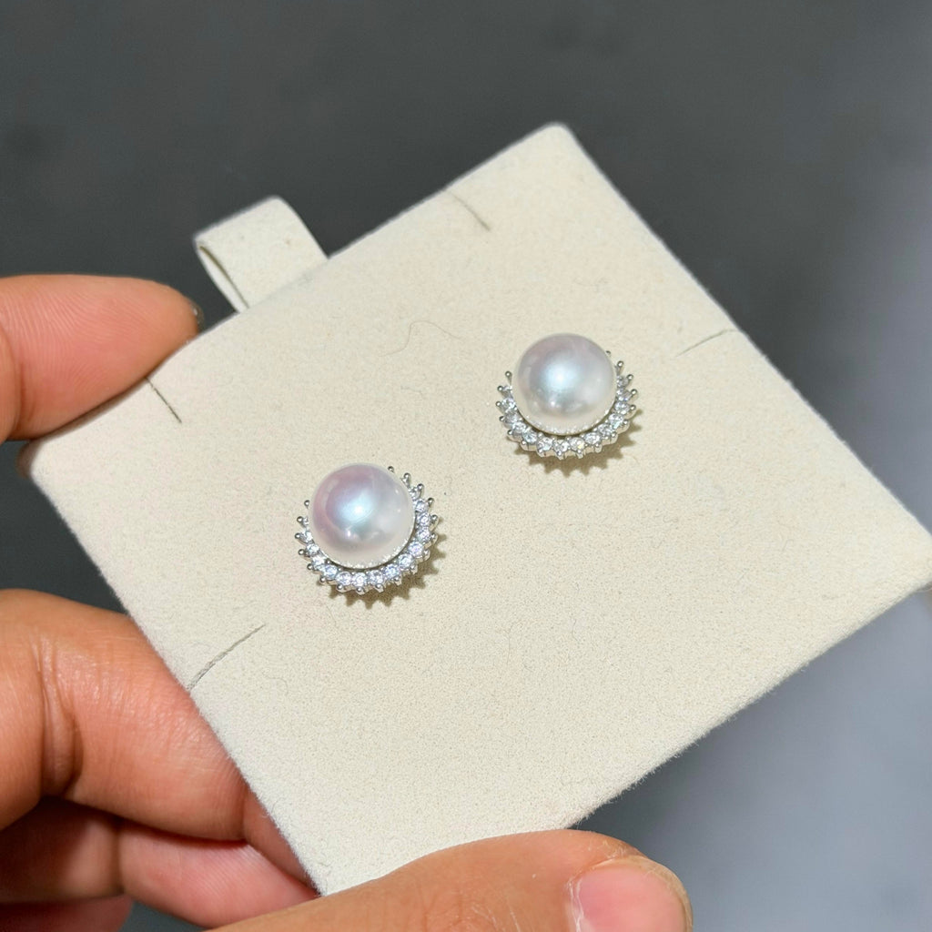 A pair of pearl stud earrings with silver crescent-shaped settings and small clear stones, displayed on a cream jewelry card held by a hand against a dark gray background.