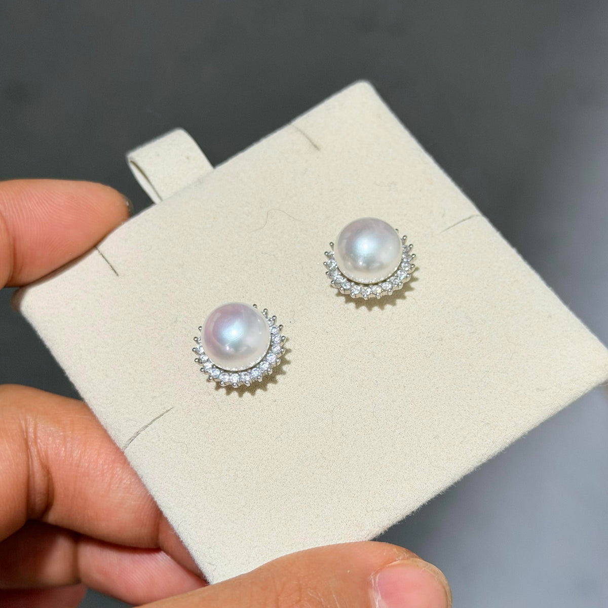 A pair of pearl stud earrings with silver crescent-shaped settings and small clear stones, displayed on a cream jewelry card held by a hand against a dark gray background.
