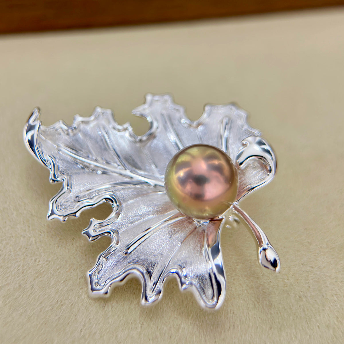 A silver leaf-shaped brooch featuring a central iridescent pearl with pink, gold, and green tones, set against a neutral beige background.