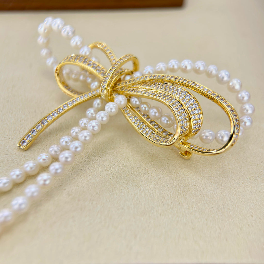 A gold bow-shaped jewelry piece with rows of clear pavé stones and draped strands of white faux pearls, displayed on a beige textured surface.