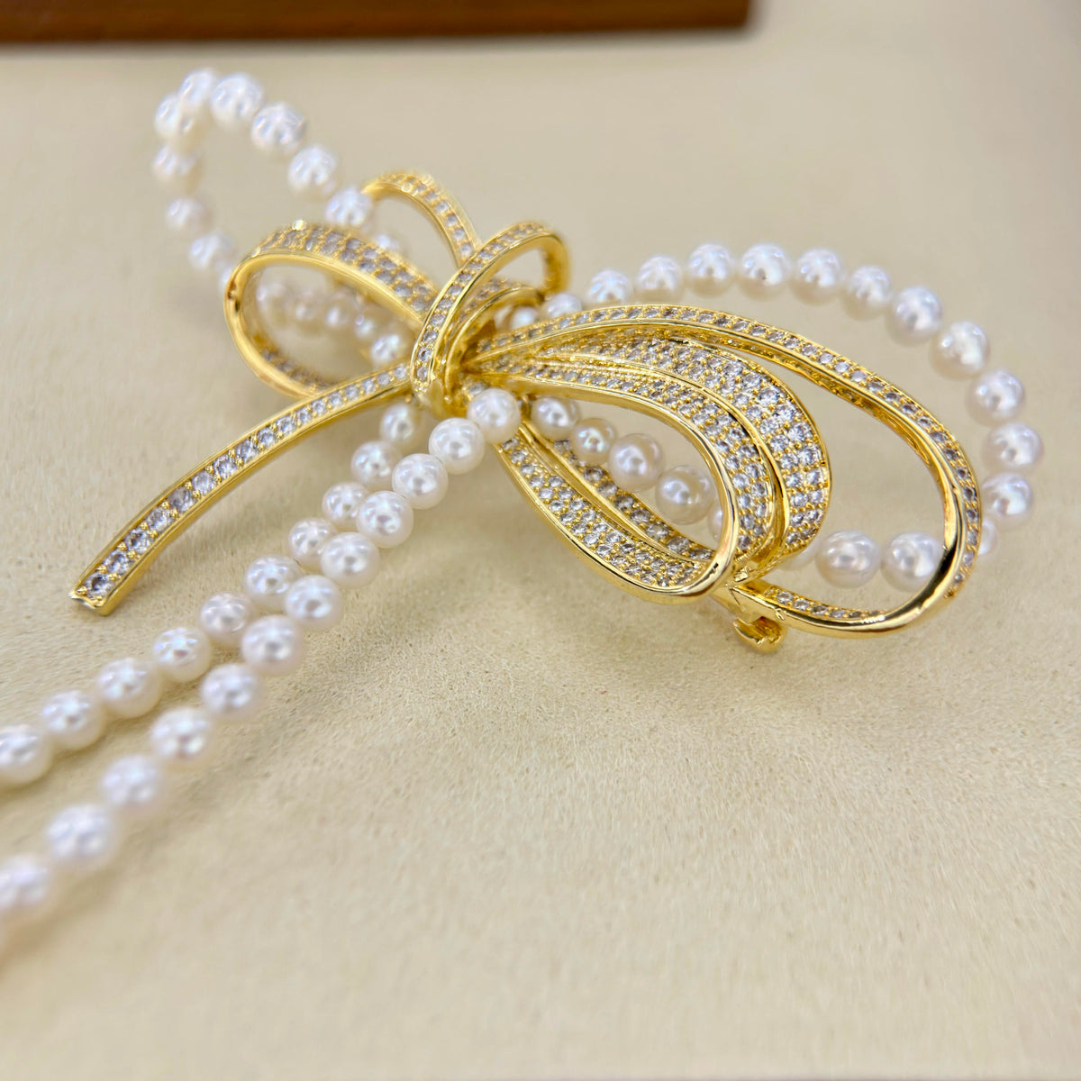 A gold bow-shaped jewelry piece with rows of clear pavé stones and draped strands of white faux pearls, displayed on a beige textured surface.