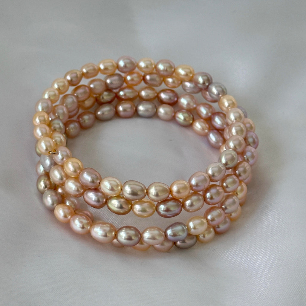 A stack of three multi-strand wrap bracelets made of irregular baroque freshwater pearls in a soft peach, pink, and champagne color, arranged on a light gray fabric background.