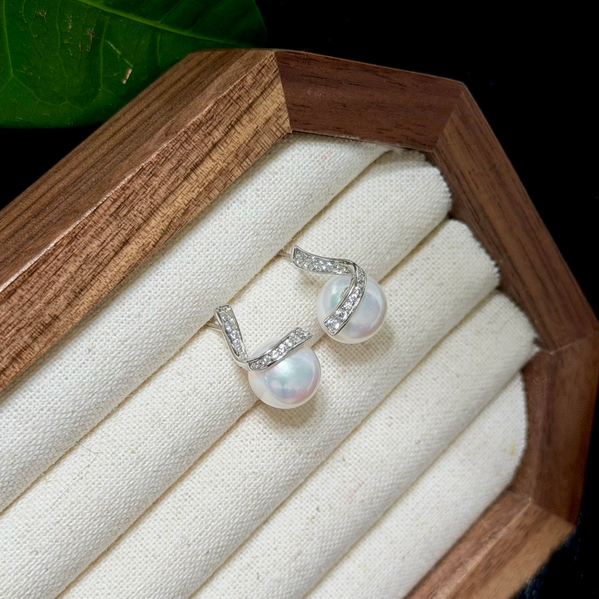 A pair of silver hoop earrings with iridescent white pearls and small clear stones, displayed on a cream fabric-lined wooden jewelry box.