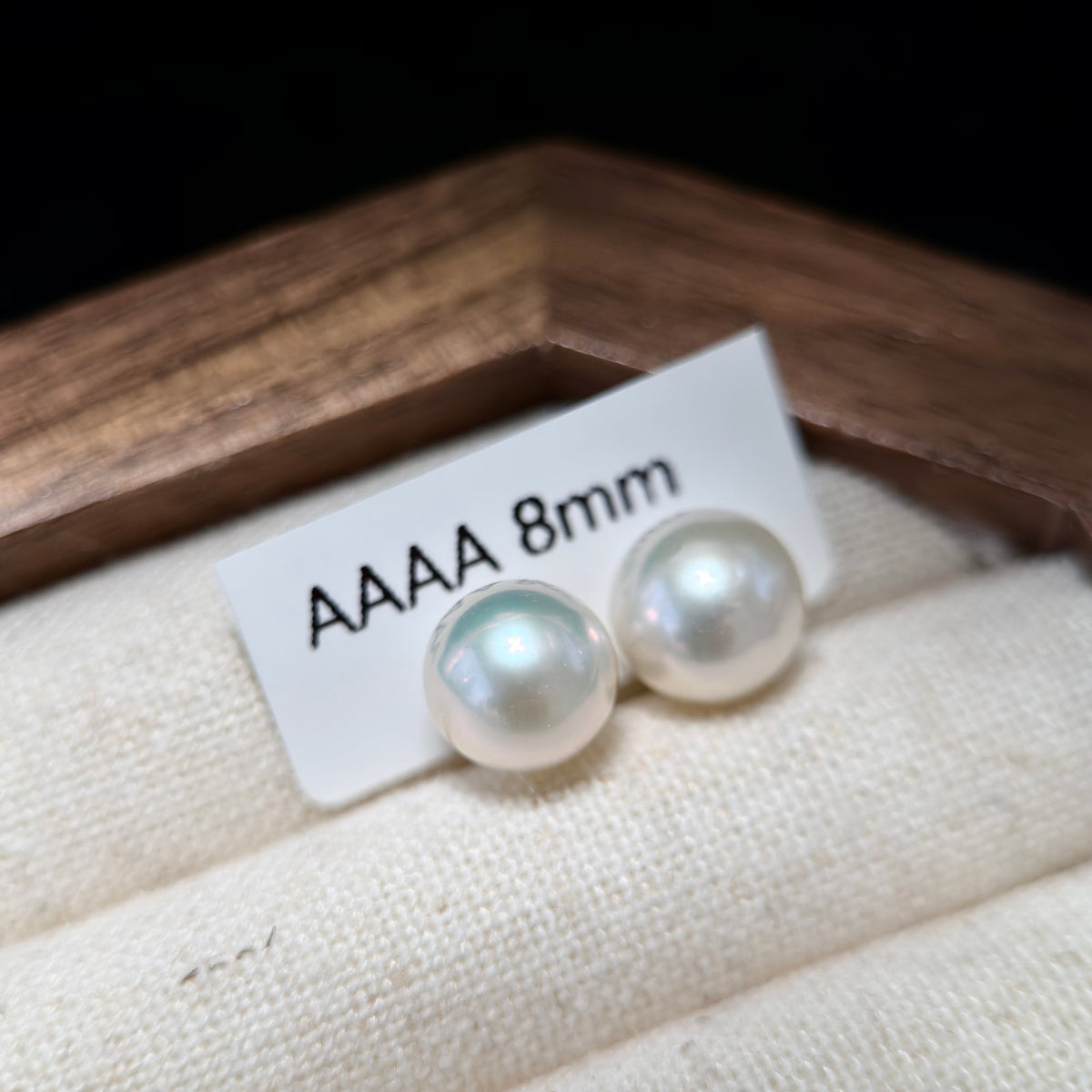Two round white pearl stud earrings displayed on a small white card labeled 'AAAA 8mm', resting on a cream fabric surface with a wooden box edge in the background.