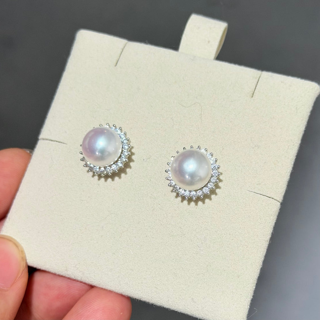 A pair of pearl stud earrings with silver settings and small clear stones, displayed on a cream jewelry card held by a hand against a dark gray background.