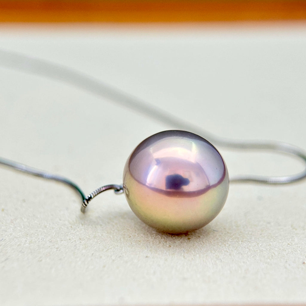 A single iridescent freshwater pearl pendant on a thin silver wire necklace, resting on a light beige textured surface.