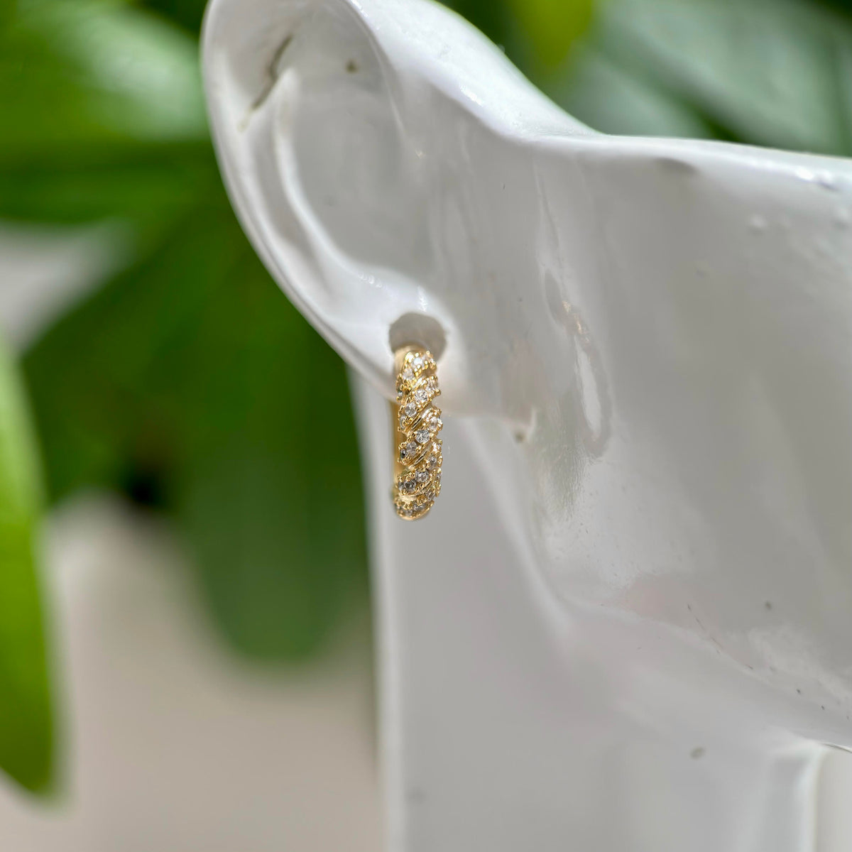 A gold huggie hoop earring with pavé-set clear stones, shown worn on a white mannequin ear with a soft-focus green plant background.