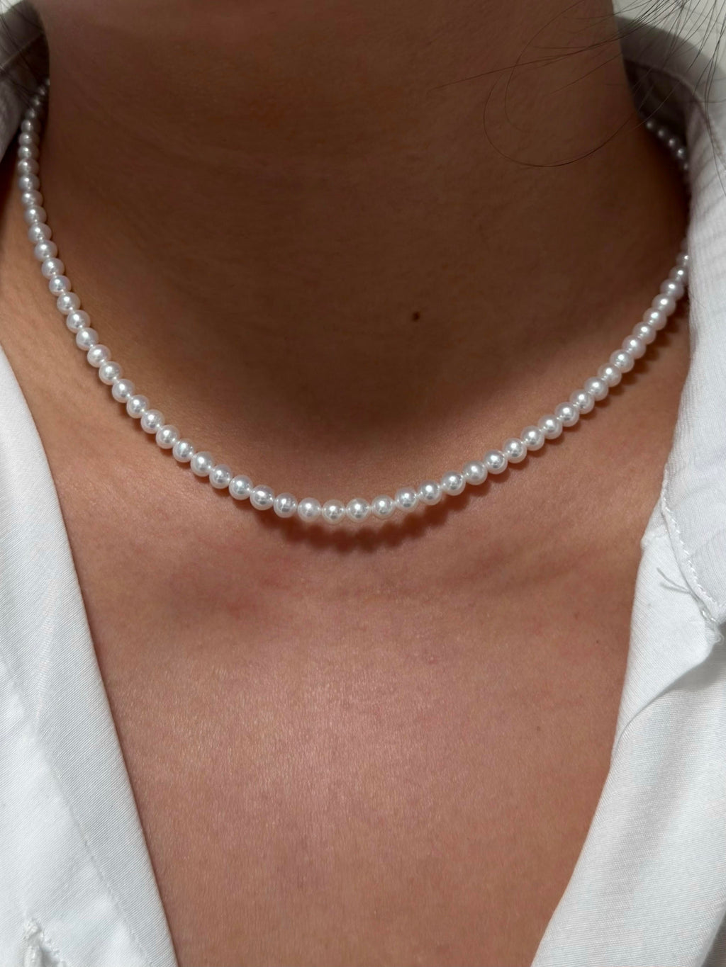 A close-up of a person wearing a classic white pearl necklace with a white collared shirt.