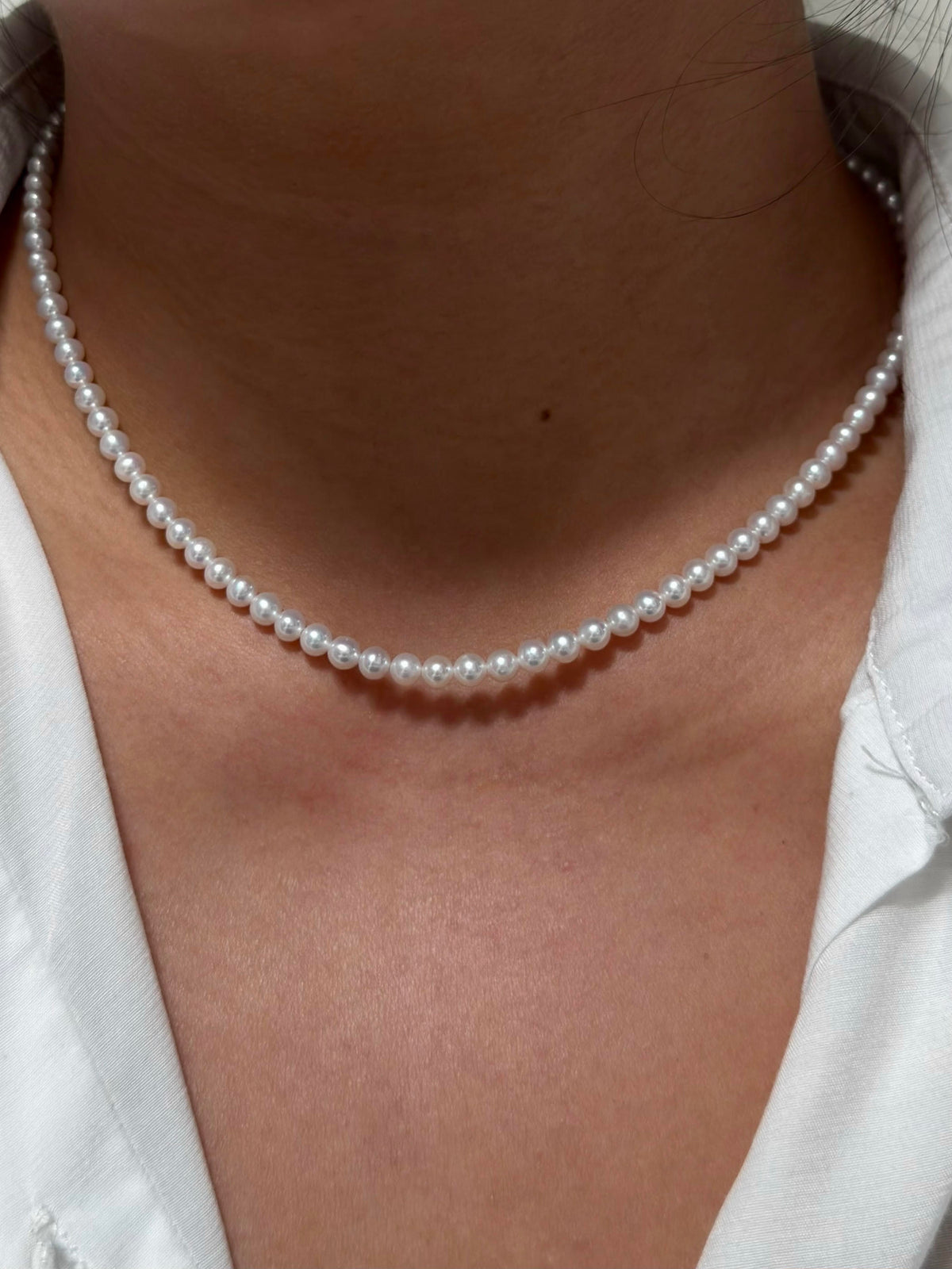 A close-up of a person wearing a classic white pearl necklace with a white collared shirt.