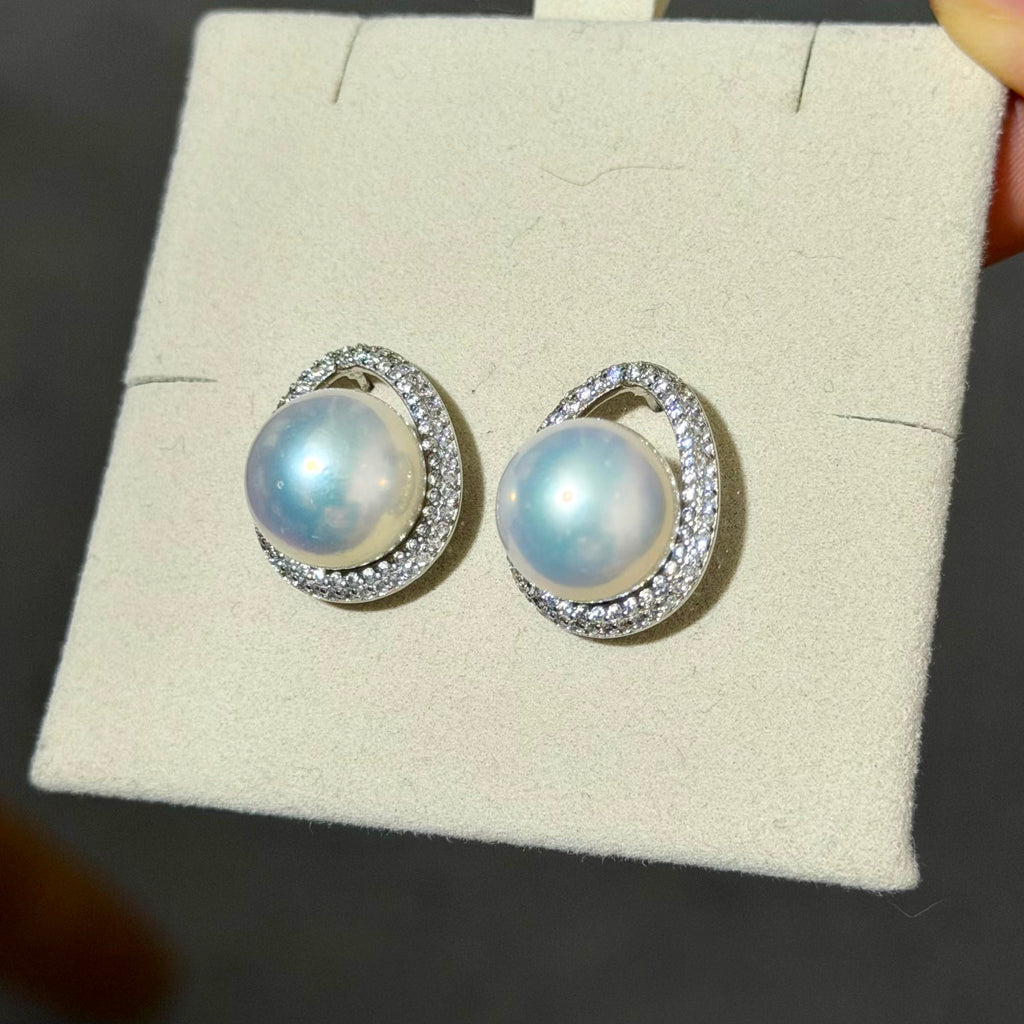 A pair of silver crescent-shaped hoop earrings with round iridescent white pearls and small clear stones, displayed on a cream jewelry card.