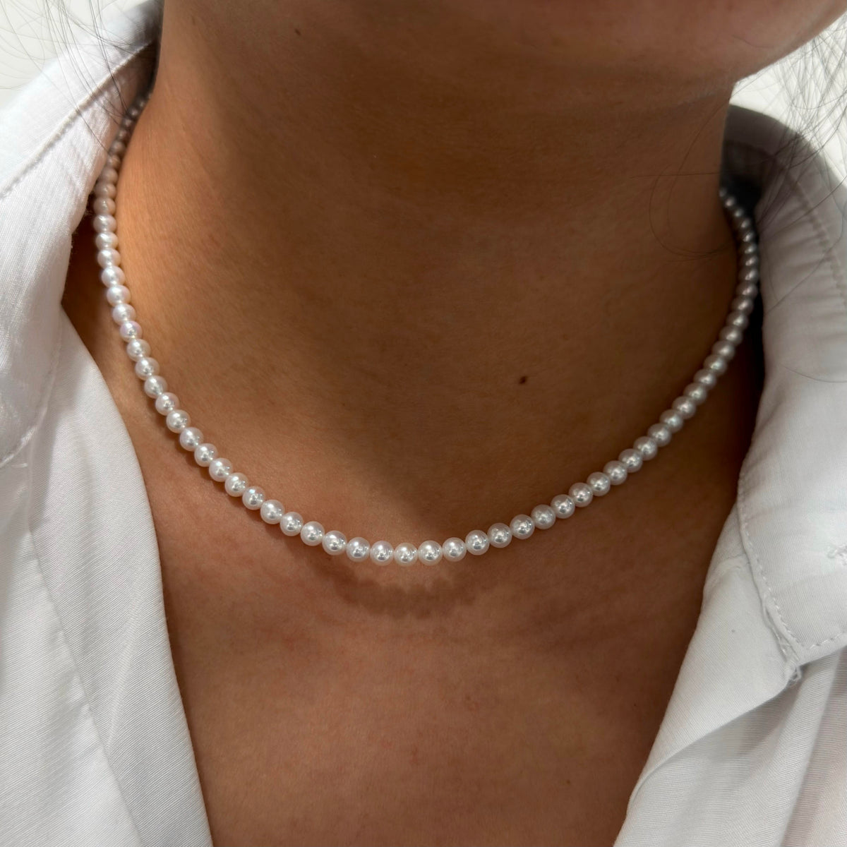 A close-up of a person's neck and collarbone area wearing a single-strand white pearl necklace over a white collared shirt.