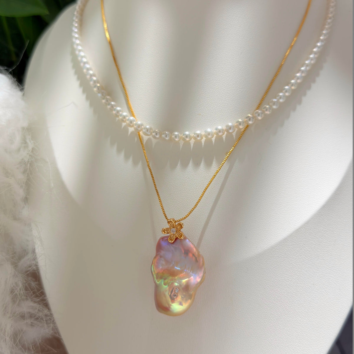 A layered necklace displayed on a white mannequin bust, featuring a delicate gold chain with a large, irregular iridescent baroque pearl pendant layered over a strand of small white pearls.