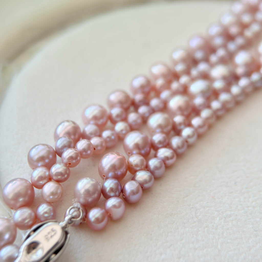 A close-up of a multi-strand pearl necklace with irregular, lustrous pink pearls and a silver clasp, displayed on a soft cream-colored surface.
