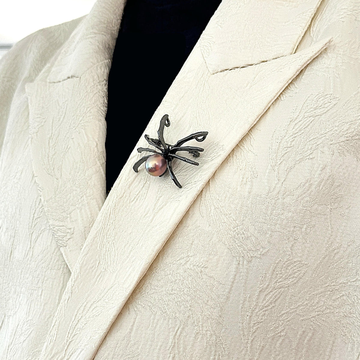 A close-up of a textured cream-colored blazer with a black spider-shaped brooch pinned to the lapel, featuring a small pink pearl at the spider's head.