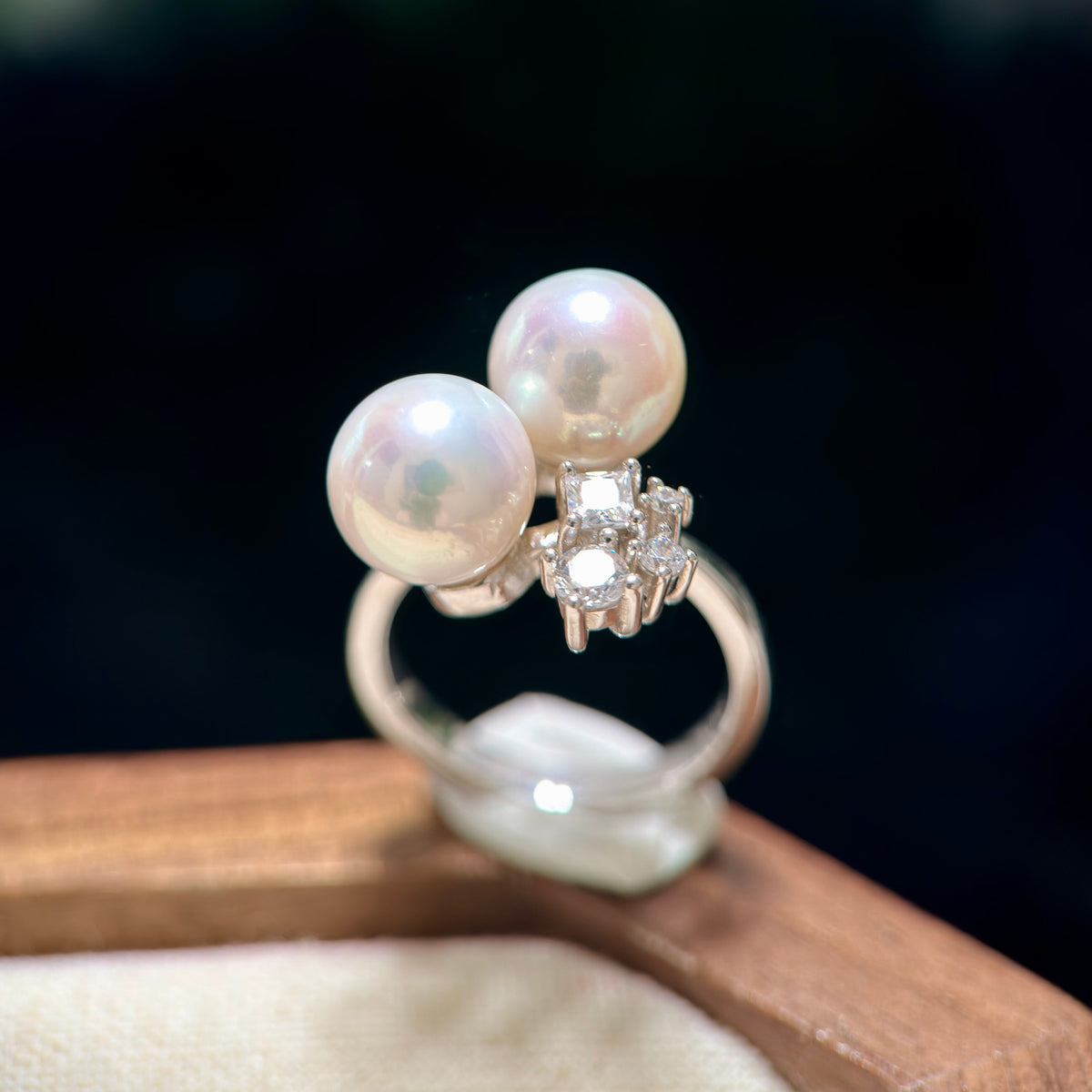 A silver ring featuring two large white pearls and a central cluster of clear gemstones, displayed on a wooden surface against a dark background.