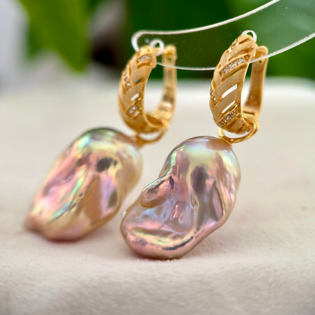 A pair of gold hoop earrings with large, organic-shaped baroque pearls in a soft pink and iridescent finish, resting on a light beige surface with a blurred green background.
