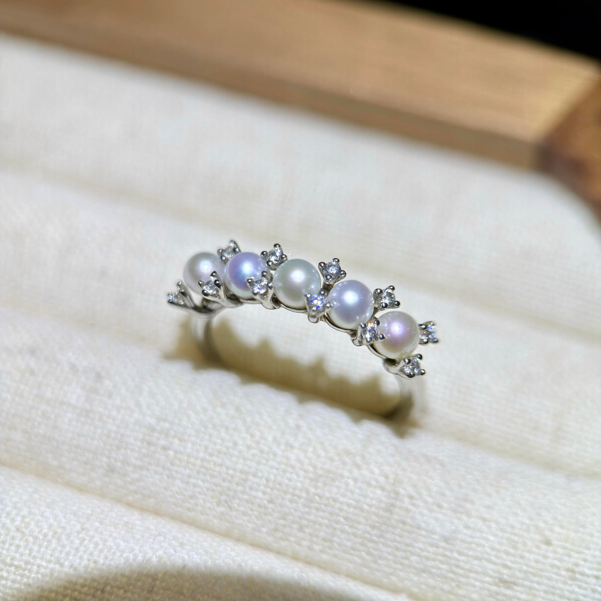 A silver ring featuring five white pearls and small clear crystals, displayed on a textured cream fabric surface.