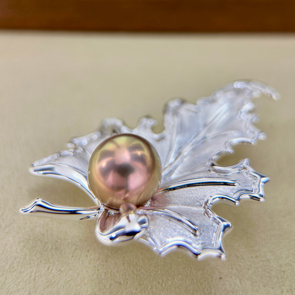 A silver maple leaf pendant brooch featuring a central iridescent pinkish pearl, displayed on a neutral beige surface.