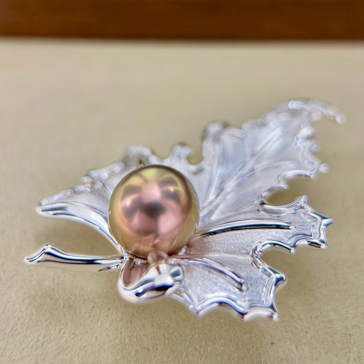 A silver maple leaf pendant brooch featuring a central iridescent pinkish pearl, displayed on a neutral beige surface.