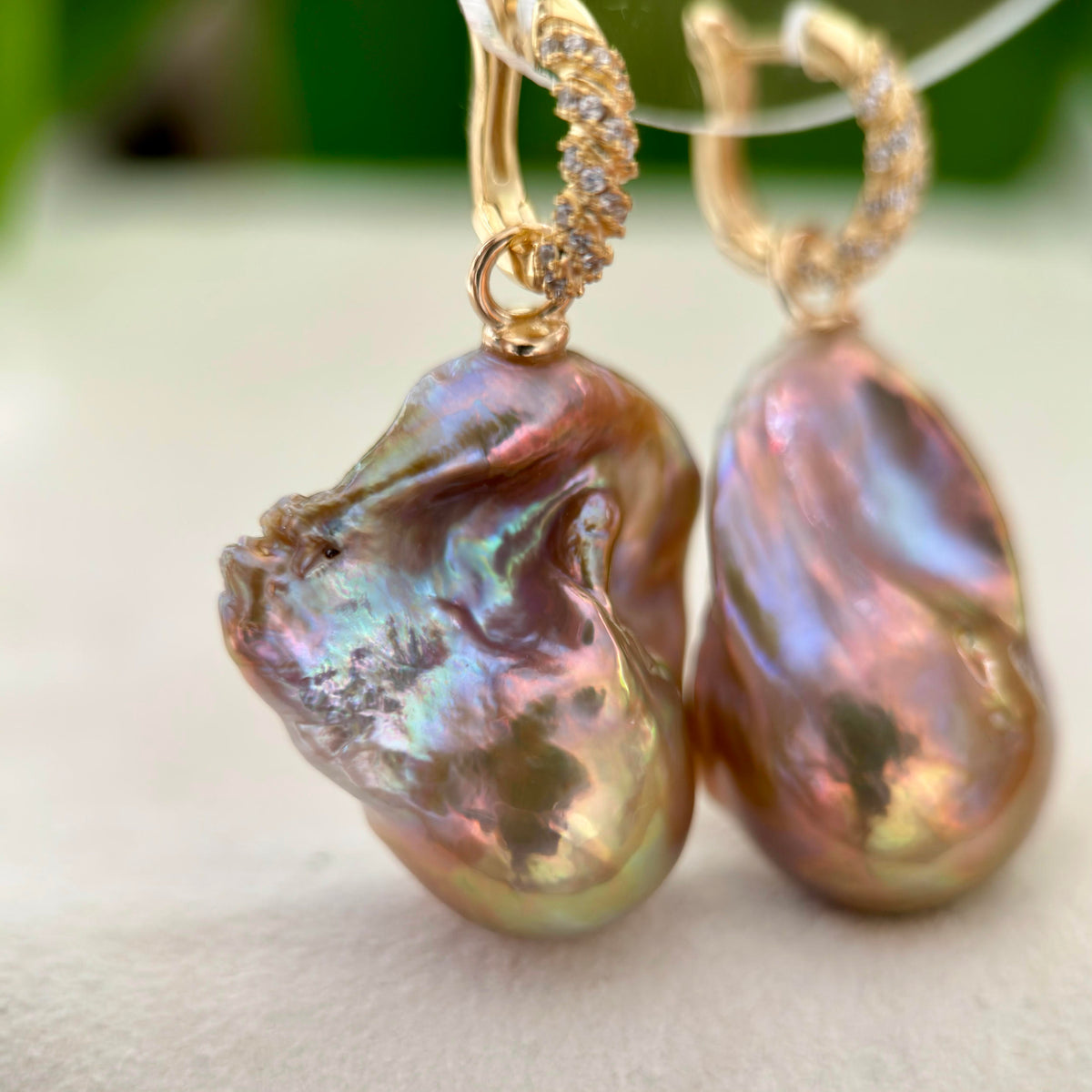 A pair of gold hoop earrings with baroque pearl drops, featuring a twisted gold top and small clear stones, resting on a light surface with a soft green background.