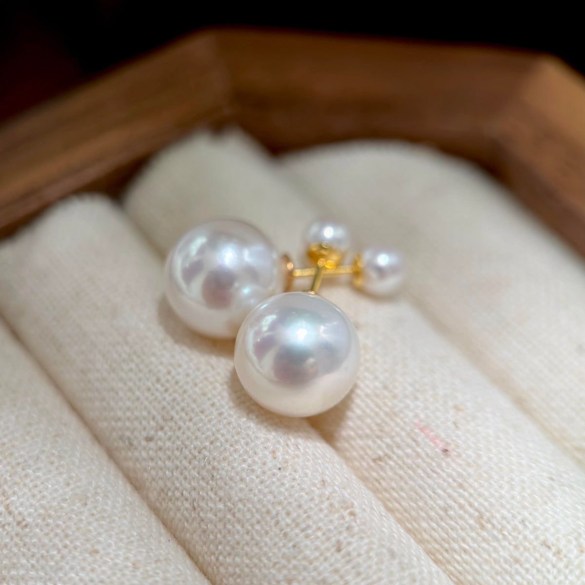 A pair of pearl stud earrings with gold posts, displayed on a cream fabric jewelry roll inside a wooden box.