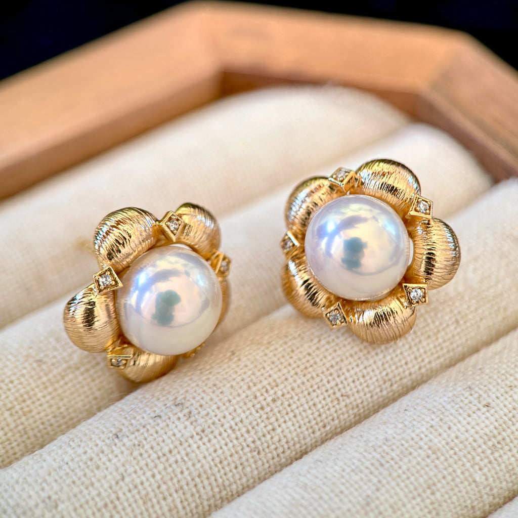 A pair of gold flower-shaped stud earrings with large white freshwater pearls and small clear accent stones, displayed on a cream fabric-lined jewelry box.