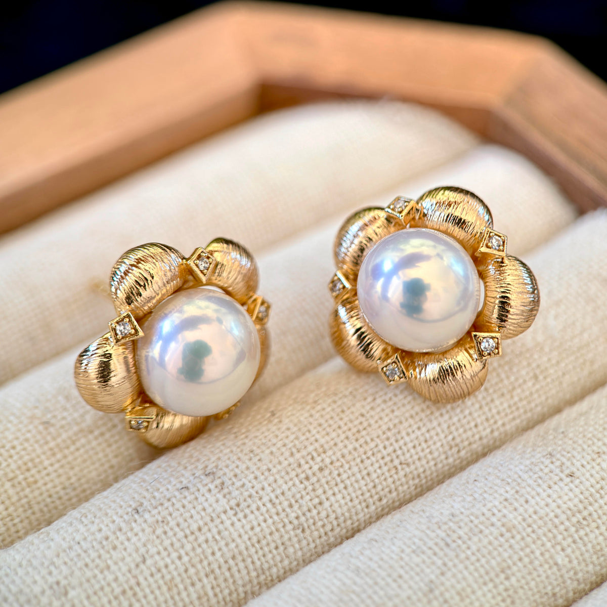 A pair of gold flower-shaped stud earrings with large white freshwater pearls and small clear accent stones, displayed on a cream fabric-lined jewelry box.