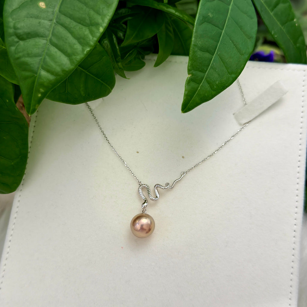 A delicate silver necklace with a small, curved silver pendant and a single dangling pinkish pearl, displayed on a white textured surface with green leaves in the background.