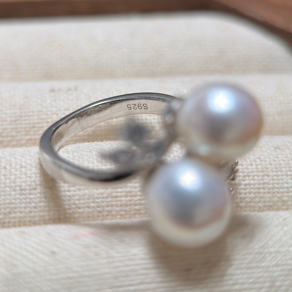 A sterling silver ring with two large white pearls, resting on a textured beige fabric surface. The inside of the band is stamped with 'S925'.