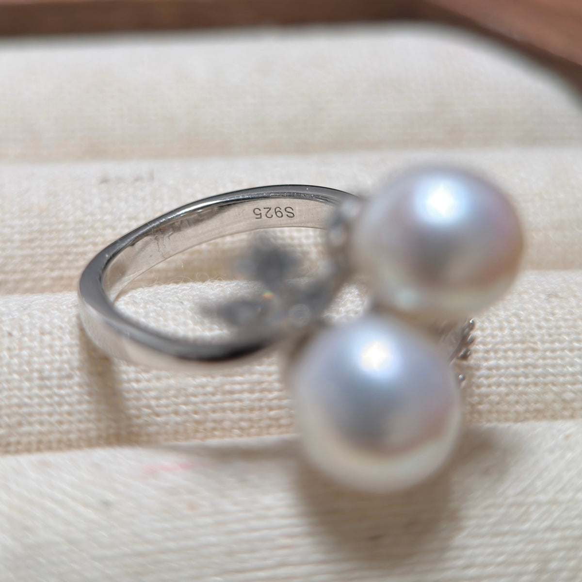 A sterling silver ring with two large white pearls, resting on a textured beige fabric surface. The inside of the band is stamped with 'S925'.