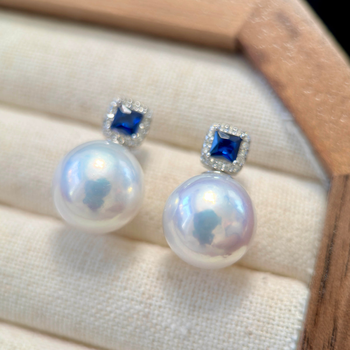 A pair of pearl stud earrings with square blue gemstones and small clear stones in a silver setting, displayed on a cream fabric jewelry box.