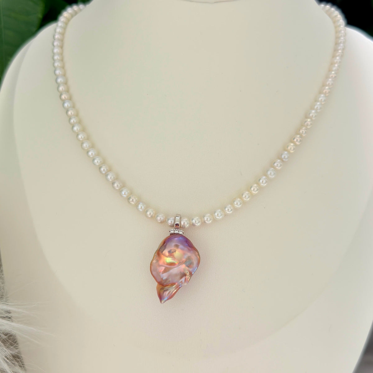 A pearl necklace displayed on a cream-colored mannequin bust, featuring a central baroque pearl pendant with iridescent pink, purple, and gold hues.